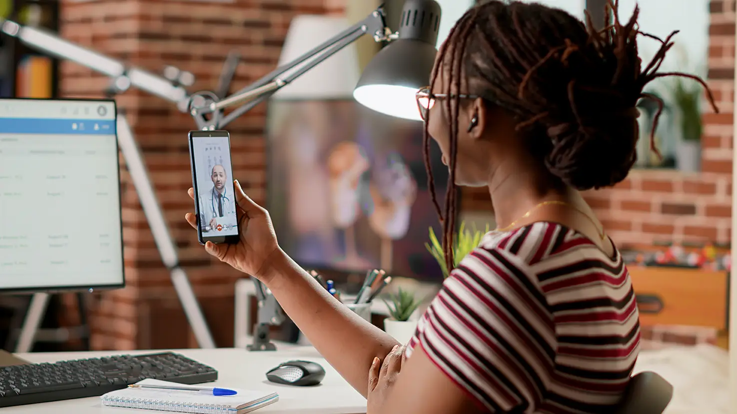 Online doctor video visit on a smartphone, with a person seated at a desk during a telehealth appointment at home.