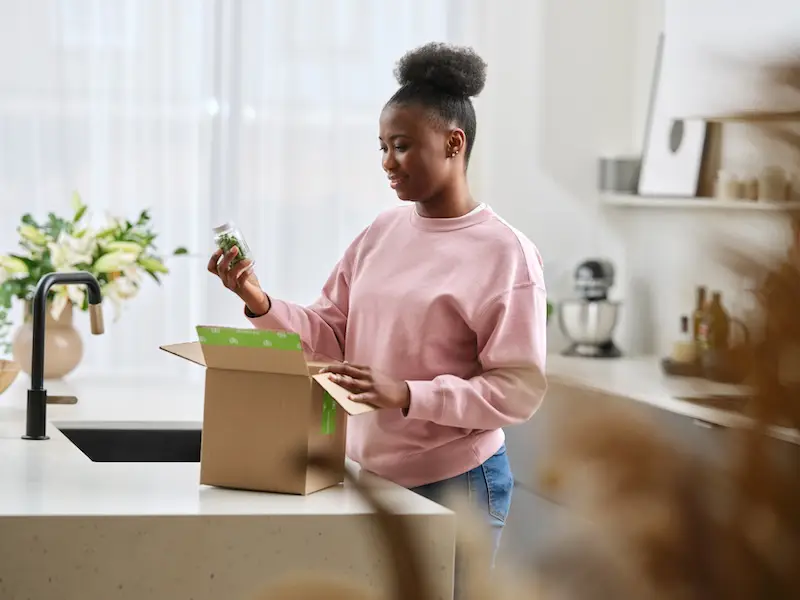 Person in a kitchen opening a package and examining a bottle of supplements, with a countertop box and home items in the background.