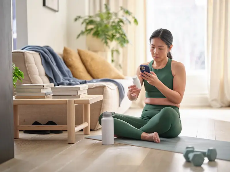 A person sits cross-legged on a yoga mat in a bright living room, looking at a smartphone while holding a small supplement container. A reusable water bottle and a pair of dumbbells rest on the floor nearby. In the background, there is a sofa with pillows and a folded blanket, a small side table with stacked books, and a potted plant, all illuminated by soft natural light from a nearby window.