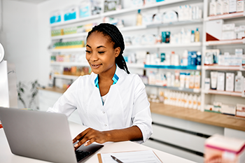Pharmacy staff member using a laptop at the counter with shelves of medications and supplies in the background
